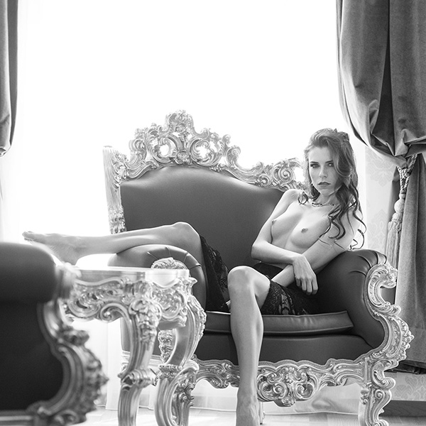 A woman in an elegant armchair, surrounded by luxurious interiors. Her expression is powerful and self-confident as she looks into the camera in a moment of calm and strength. A perfect representation of contemporary nude photography.