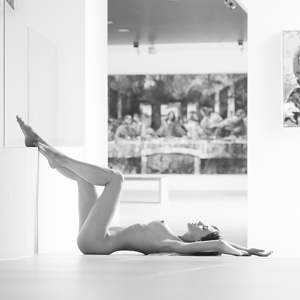 Black and white shot of a woman in artistic nude photography lying on the floor in a modern room with artwork in the background