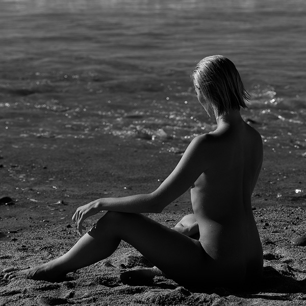 Erotische Aktfotografie: Eine nackte Frau sitzt im Sand, den Blick auf das Meer gerichtet, während die Wellen brechen.