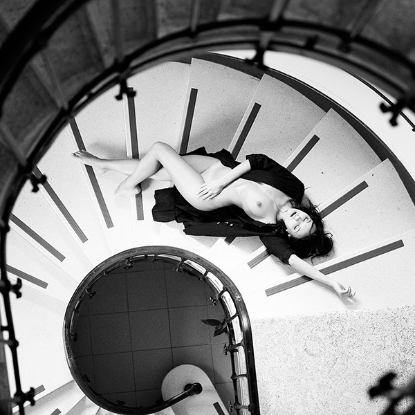 Erotische Fotografie einer Frau in schwarzem Hemd, die sich auf einer Treppe im minimalistischen Raum liegend positioniert. Ihre Körperhaltung strahlt Selbstbewusstsein und Eleganz aus.