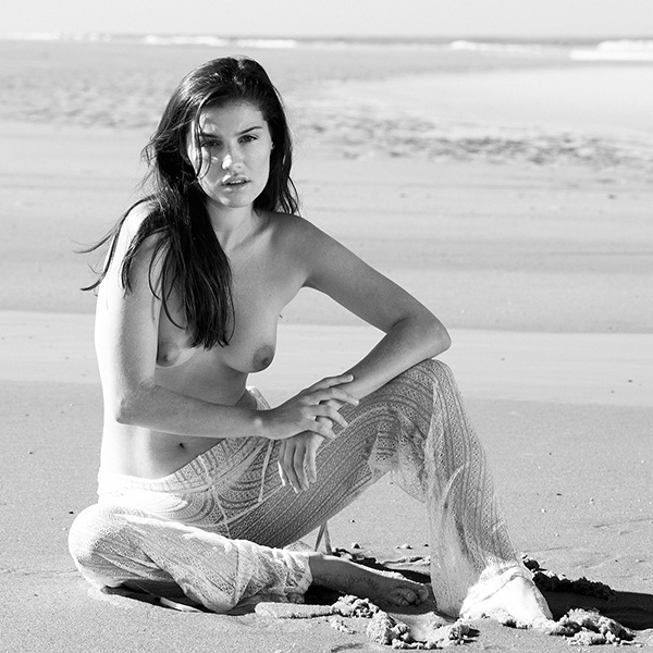 Erotic photograph of a woman sitting in the sand on the beach, a thoughtful gaze directed into the distance.