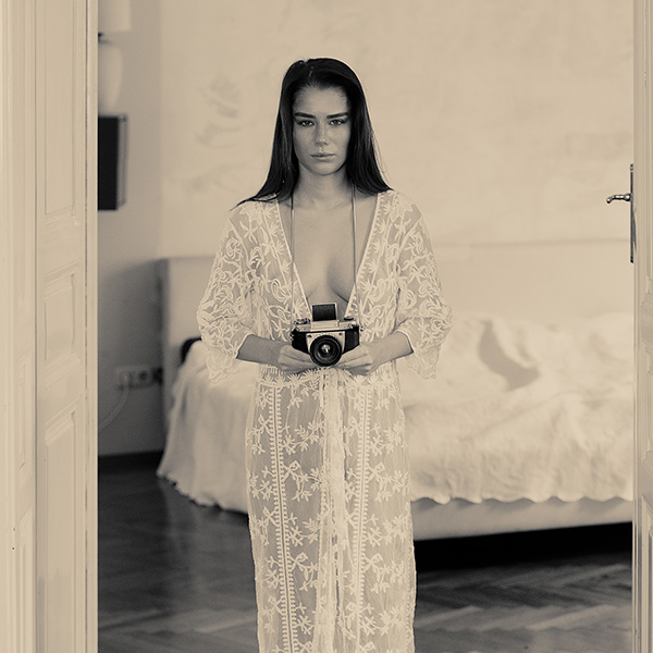 Woman in white wardrobe with camera in hand in front of neutral background - artistic nude photography.