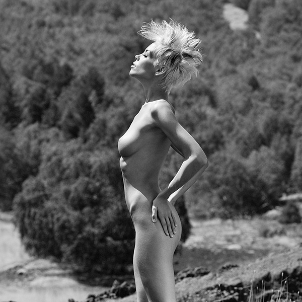 Eine Frau steht nackt in der Natur, der Wind in den Haaren – erotische Aktfotografie voller Bewegung und Kraft.