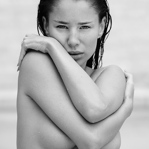 Erotische Aktfotografie einer Frau mit nassen Haaren, die sich selbst umarmt, mit intensivem Blick und Wassertropfen auf der Haut.