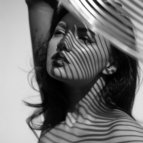 Erotic photograph of a woman covered by abstract shadow structures of narrow, vertical lines that appear on her body and face. Her pose is pensive and sensual as she looks slightly to the side. The dramatic contrast between light and shadow highlights her contours and emphasizes the strong visual impact of this black and white image.