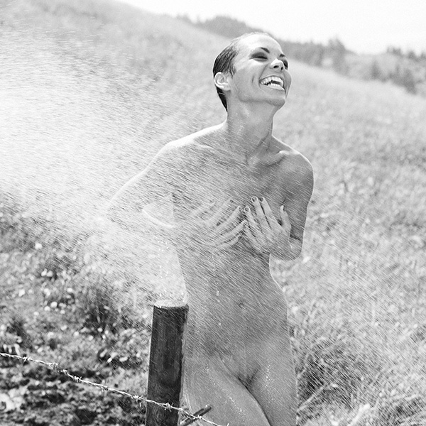 Erotische Aktfotografie einer nackten Frau unter einem Wasserstrahl, der sie im Outback umgibt, während sie sich von Dreck befreit. Ein Moment der Freiheit und der Lust.