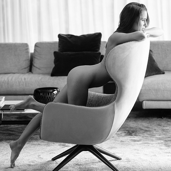 Erotic photograph of a woman in a relaxed position on an armchair, surrounded by minimalist design and subtle lighting.